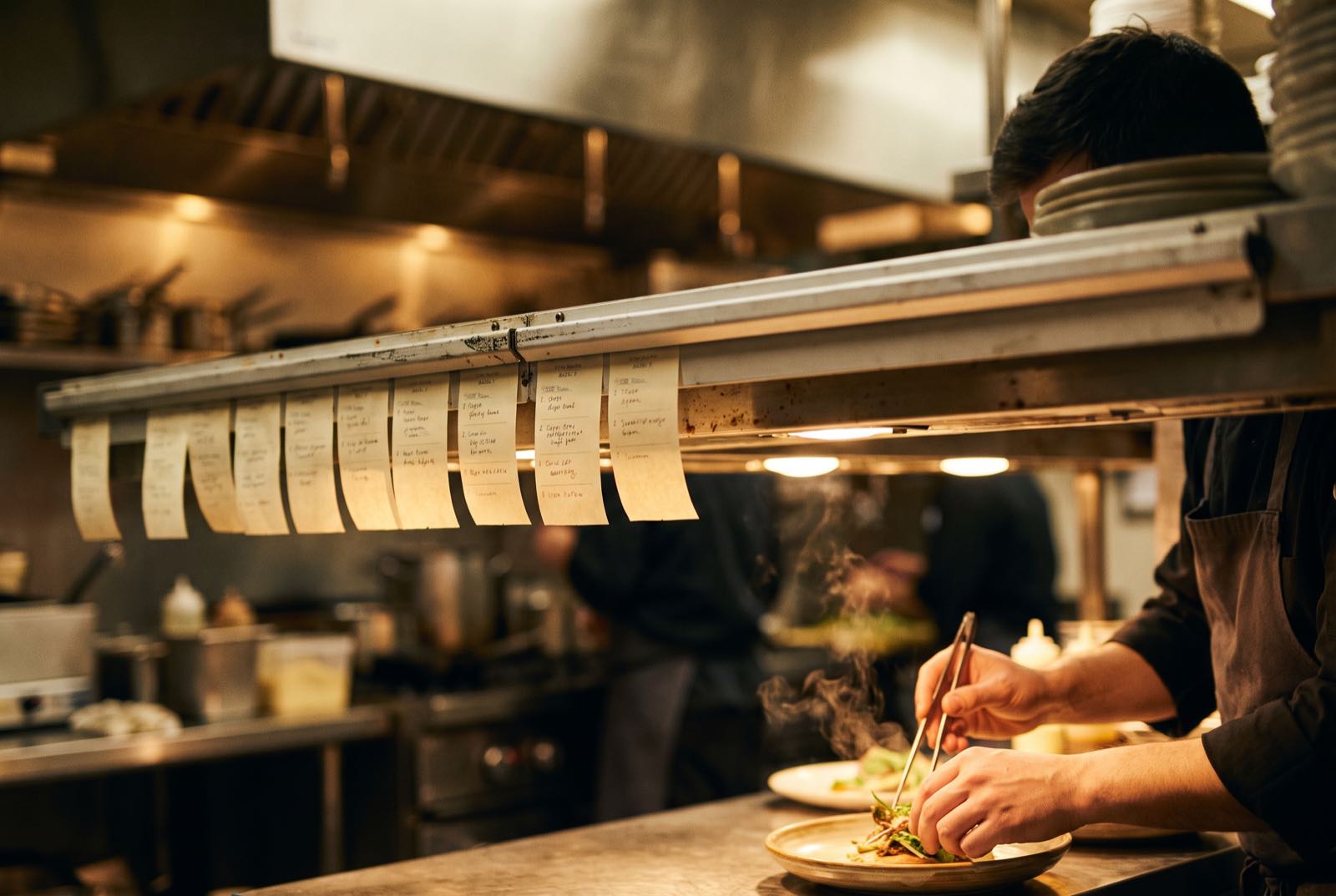 One unbroken line of order tickets on the pass while a chef plates a dish